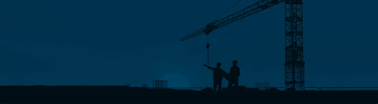 Silhouettes of two construction workers on a building site with a large crane in the background at dusk.