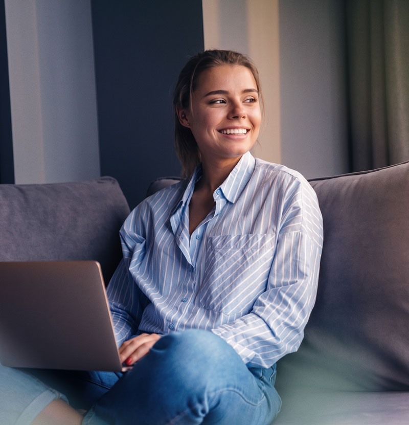 Image of a person sitting on their sofa holding a laptop while looking aside and smiling.