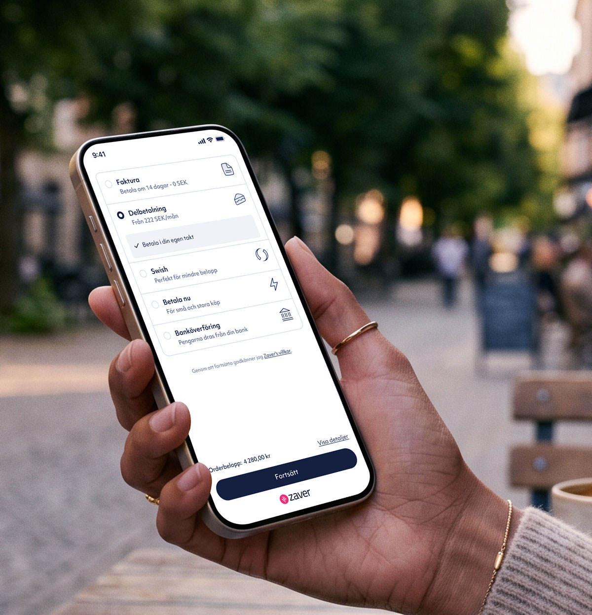 Hand holding smartphone showing the Zaver checkout displaying payment options like invoice, installment, Swish, and bank transfer.