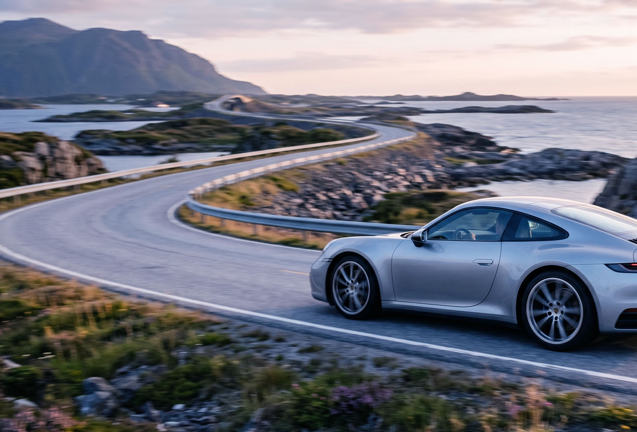 Silver car driving on a winding coastal road with mountains and ocean in the background.