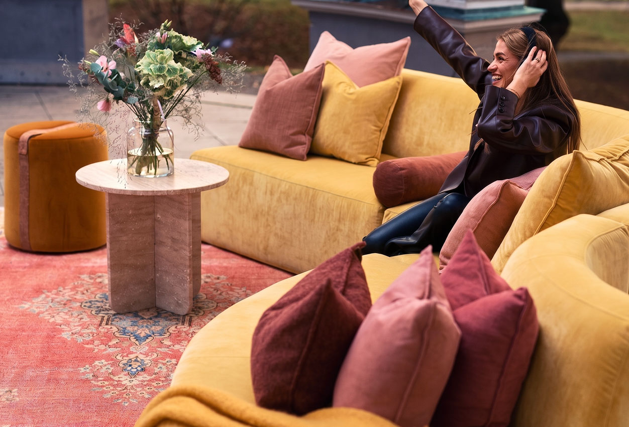 Smiling woman sitting on a yellow couch with pink and brown pillows in a cozy outdoor setting.