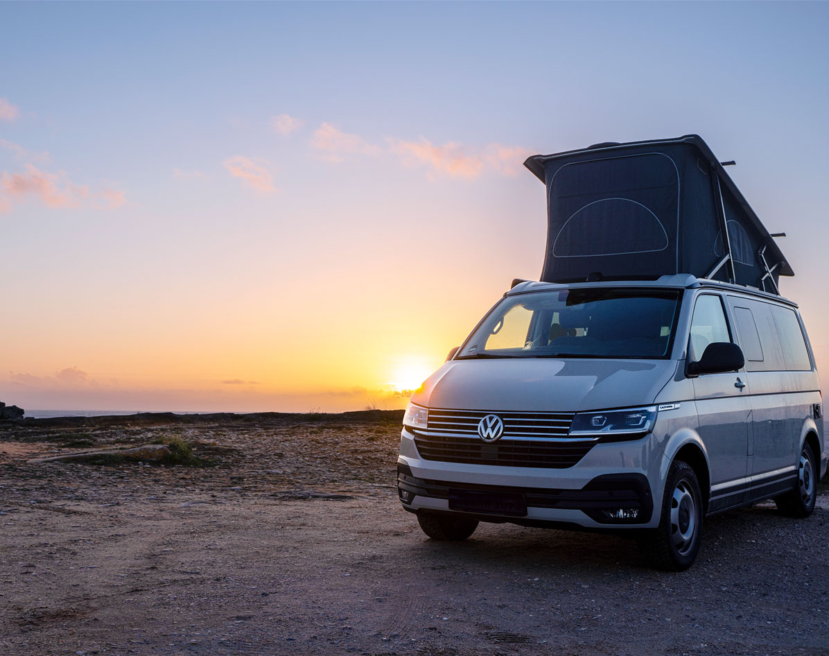 White camper van with roof tent parked on rocky ground at sunset with clear sky.