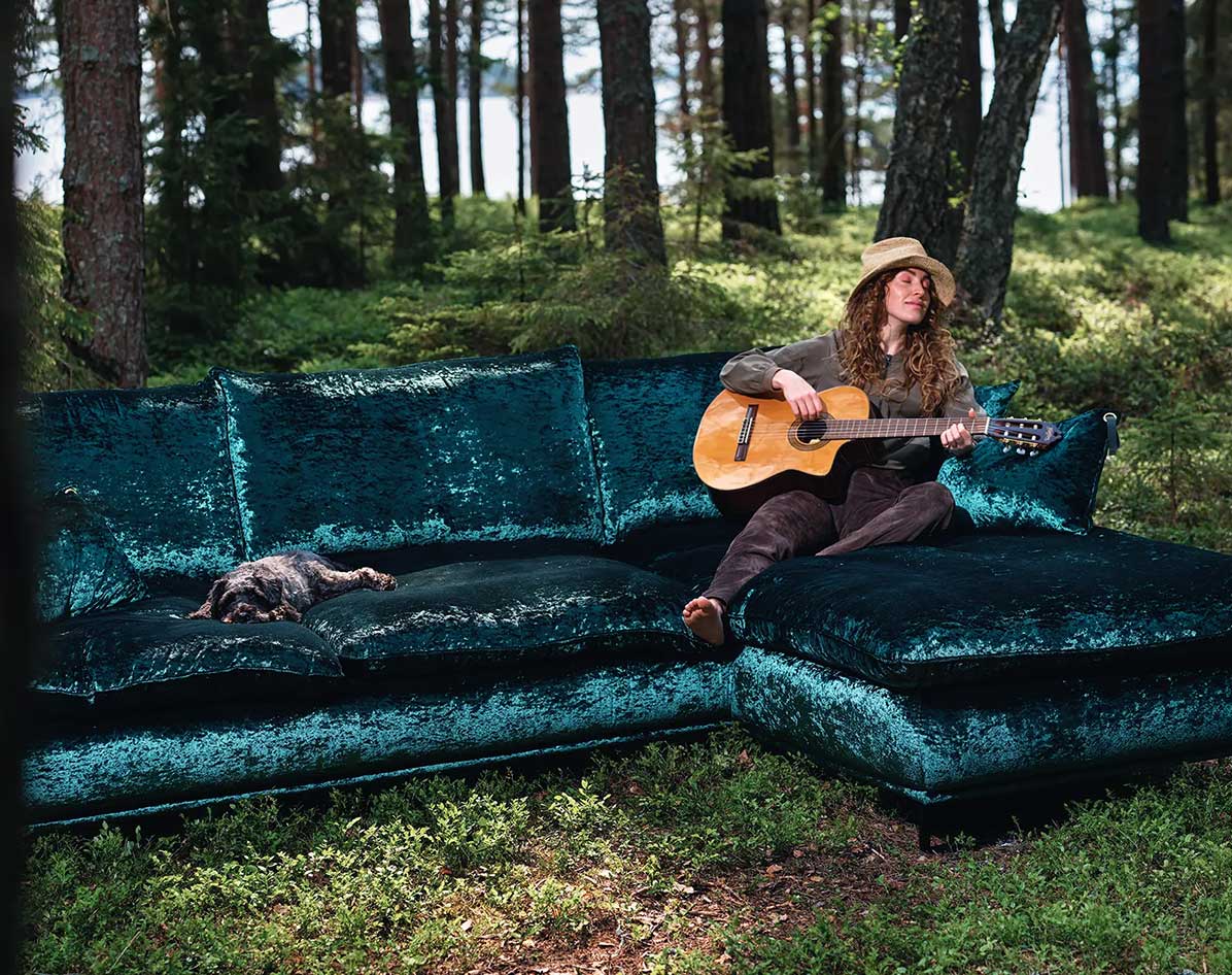 Woman in a hat plays guitar on a teal velvet sofa with a dog lying next to her in a forest.