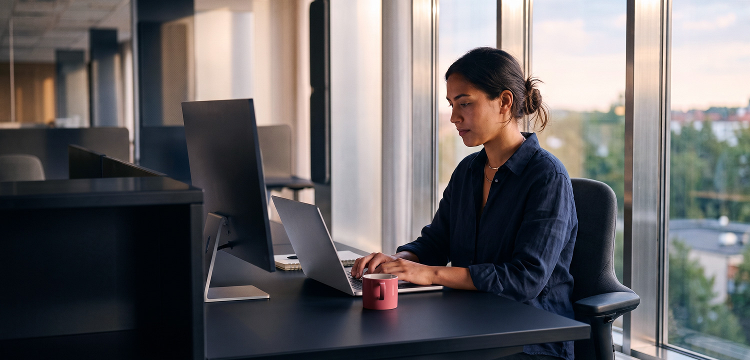 Woman working on a laptop at a desk in an office with large windows and a pink coffee mug.