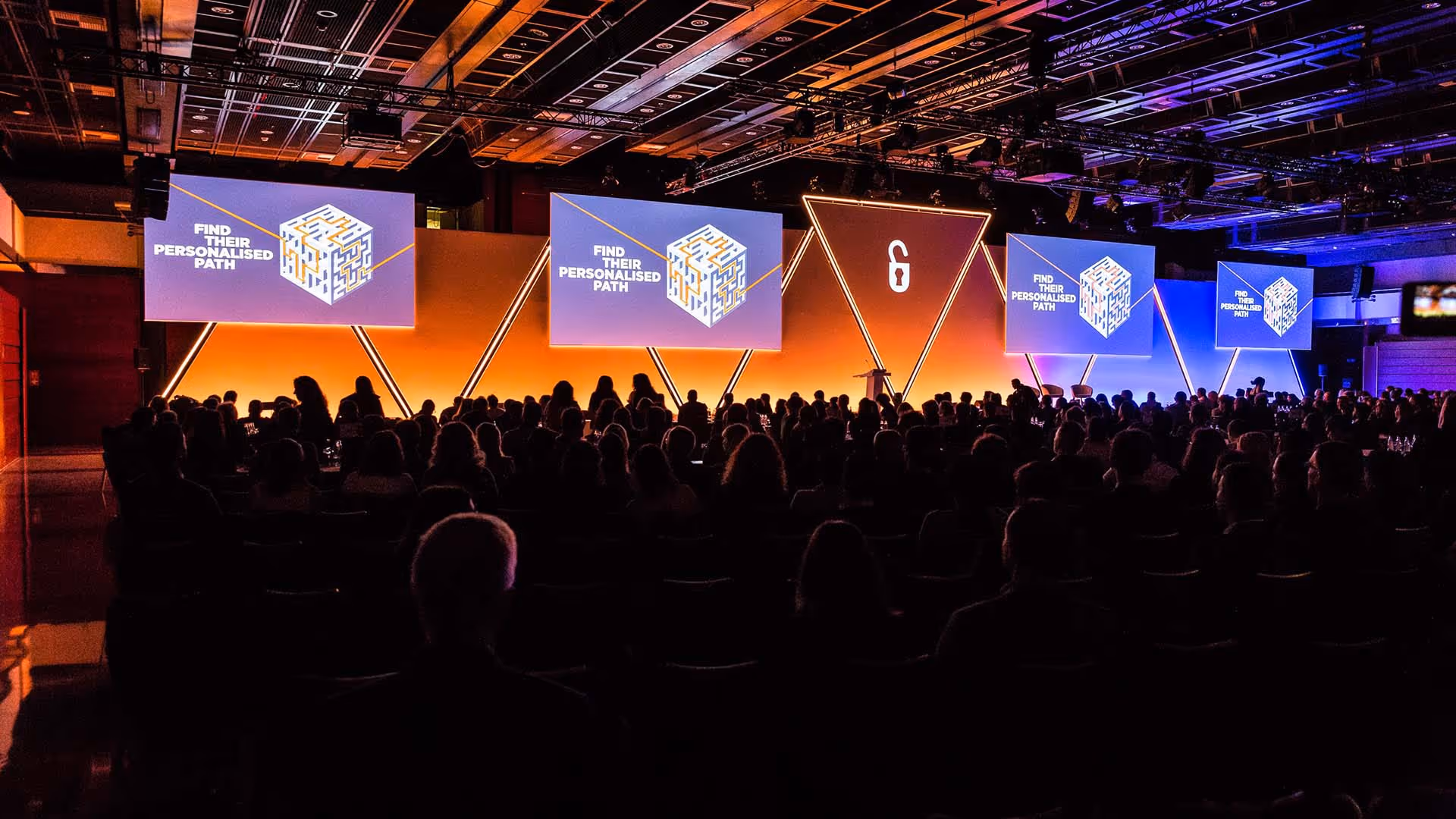 Audience look at dramatic stage with six screens and podium