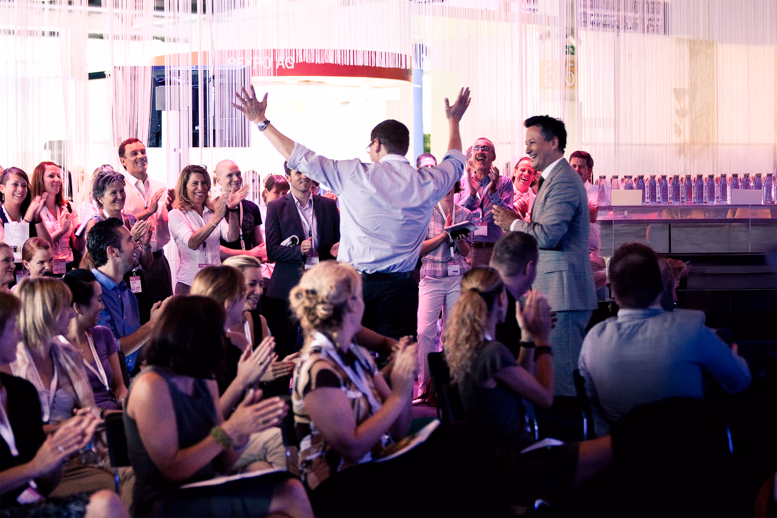 Applauding corporate event audience standing around speaker with raised arms.