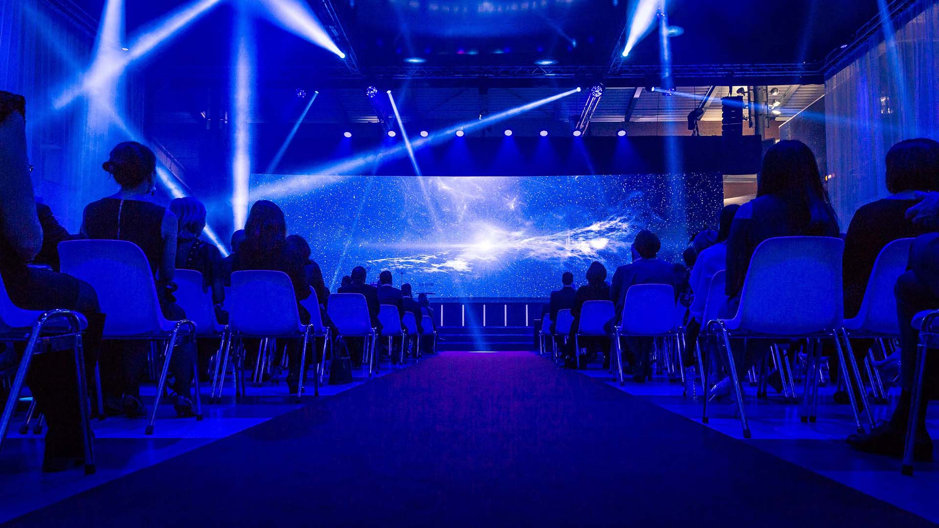 Audience seated in blue-lit meeting room with dramatic image on screen.