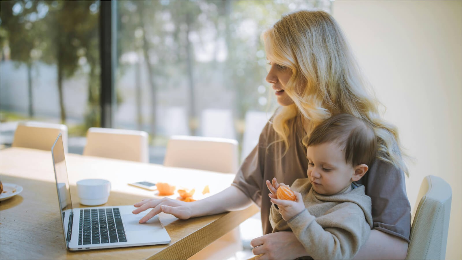 mom sat working at a laptop in her kitchen with a child sat on her knee