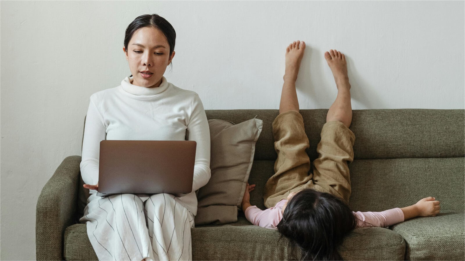 mom working on a laptop sat on the sofa with a kid playing next to her 