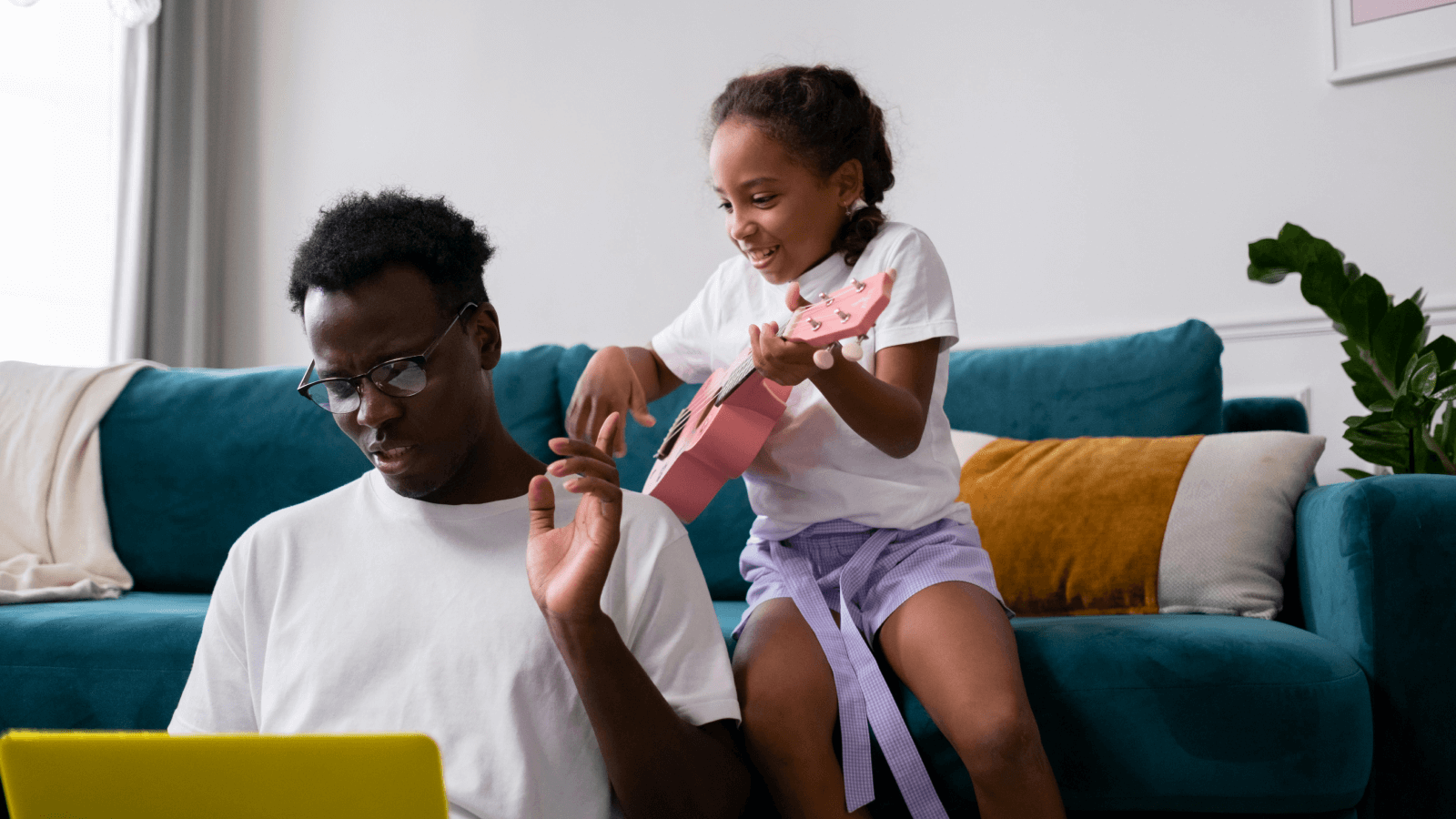 dad working from home with his daughter sat on the sofa playing a toy guitar