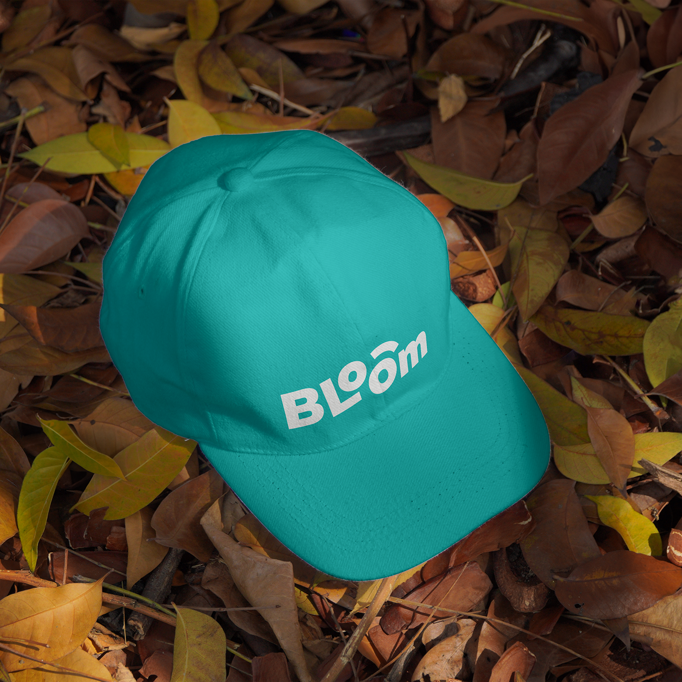 A teal baseball cap with the Bloom logo embroidered on the front, placed on autumn leaves.