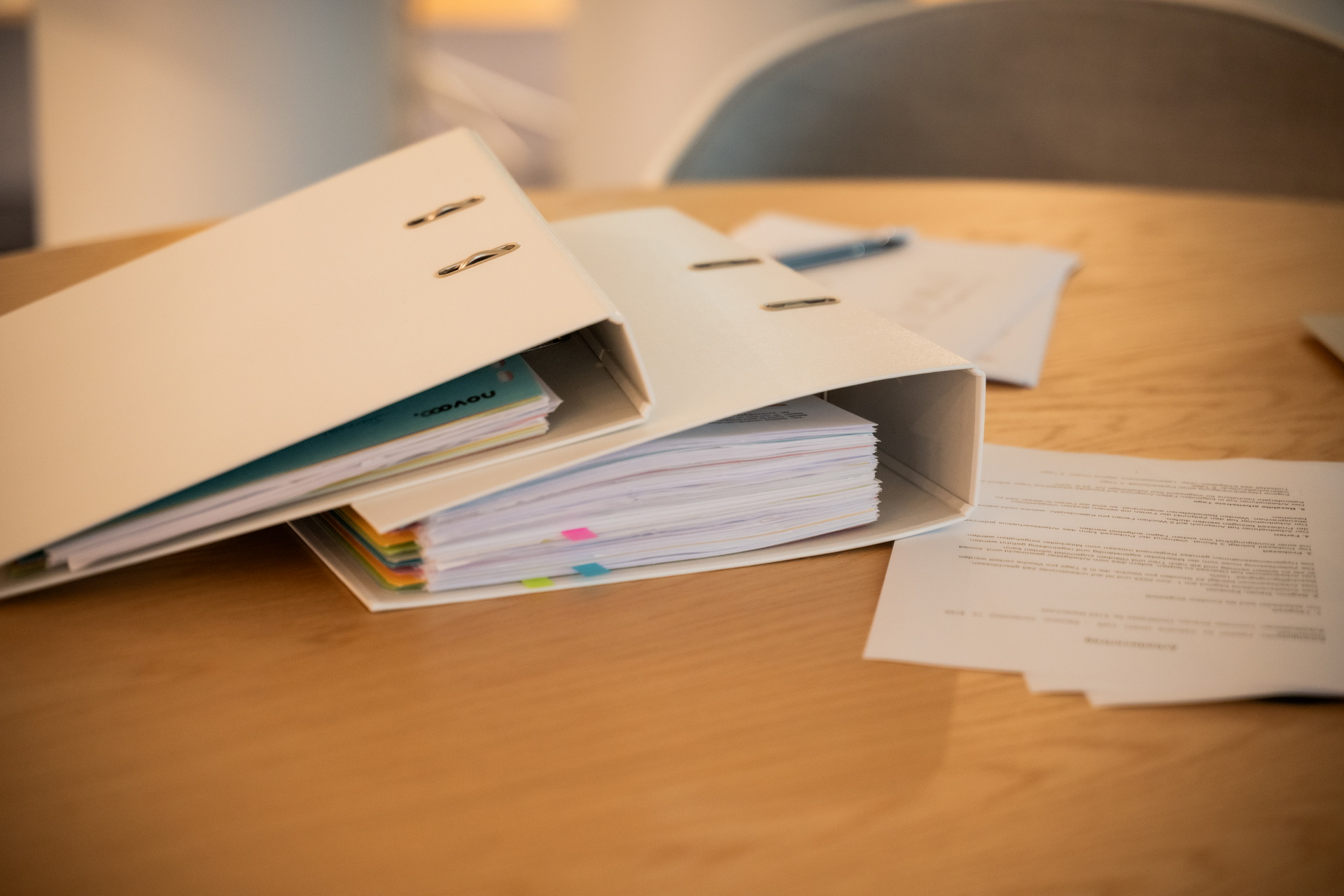 Zwei weiße Ordner mit vielen Dokumenten und farbigen Trennblättern auf einem Holztisch, daneben liegen weitere Papiere und ein Stift - ein Symbol für Buchhaltung und Jahresabschluss.