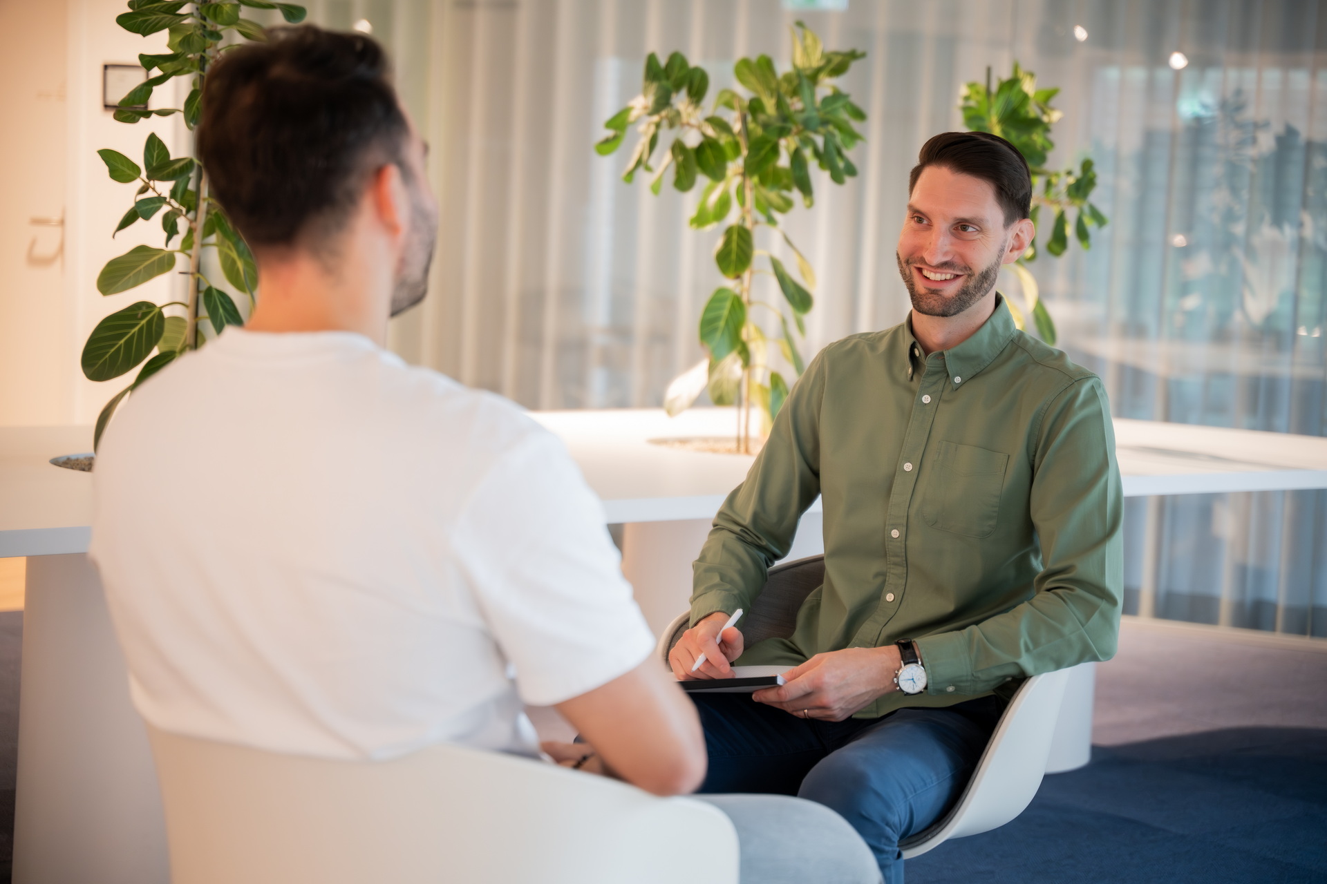 Zwei Männer im Gespräch in einem modernen Büro, einer notiert etwas auf einem Block - ein Symbol für Personal- und Businesscoaching.