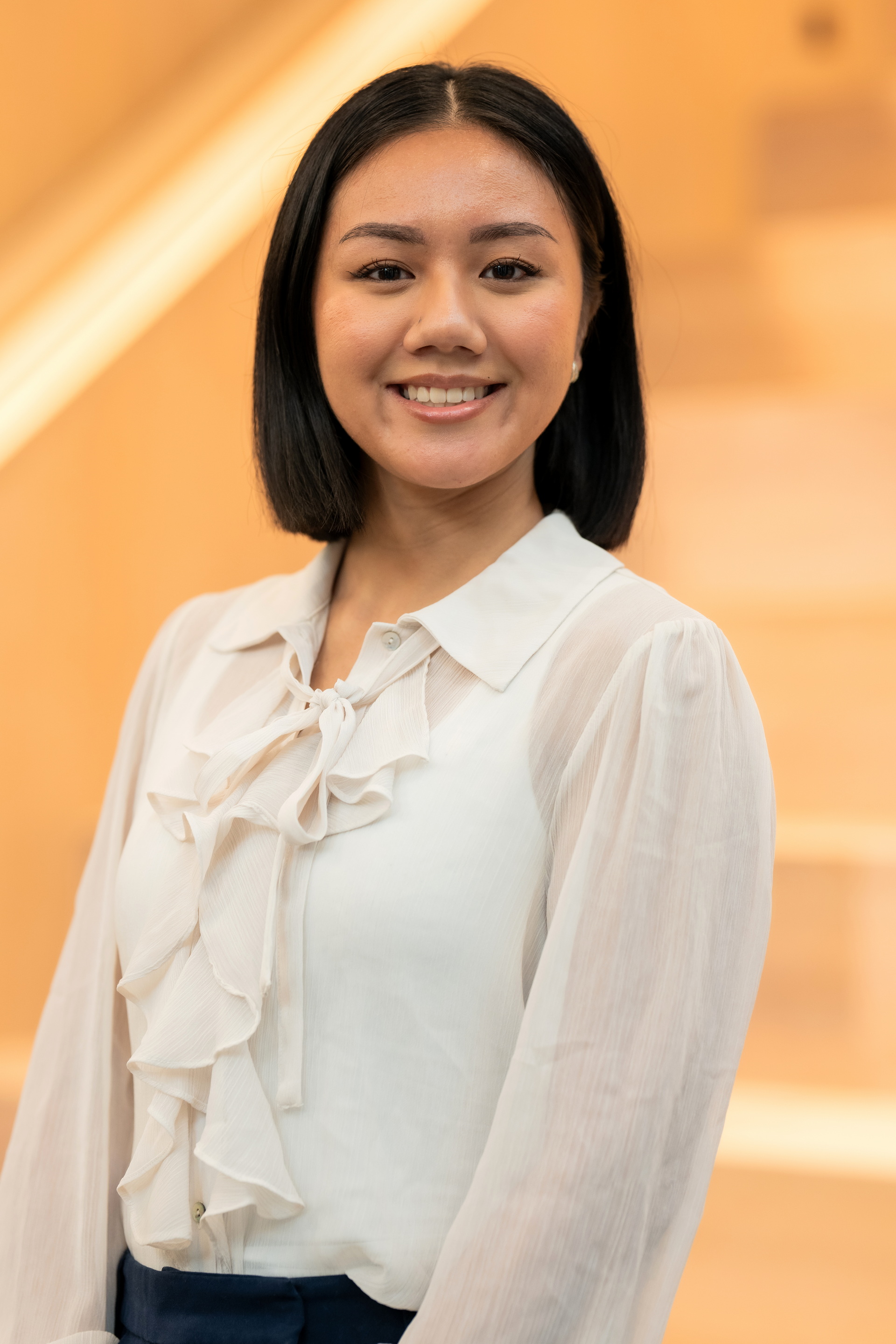 Porträt einer lächelnden jungen Frau mit schwarzen, schulterlangen Haaren und weißer Bluse mit Rüschen vor unscharfem, warmem Hintergrund.