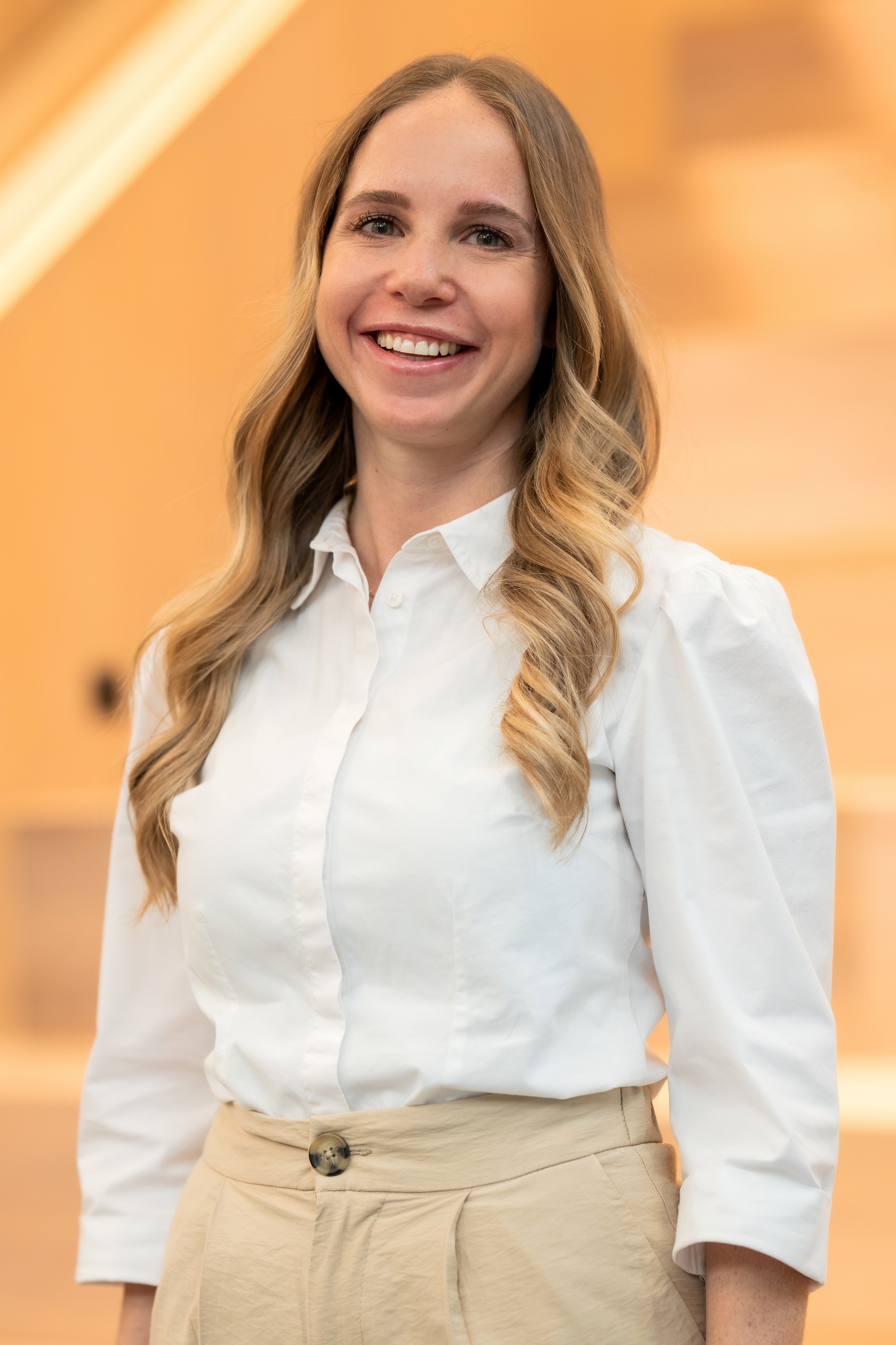 Lächelnde junge Frau mit langen, gewellten blonden Haaren trägt weiße Bluse und beige Hose vor hellgelbem Hintergrund.