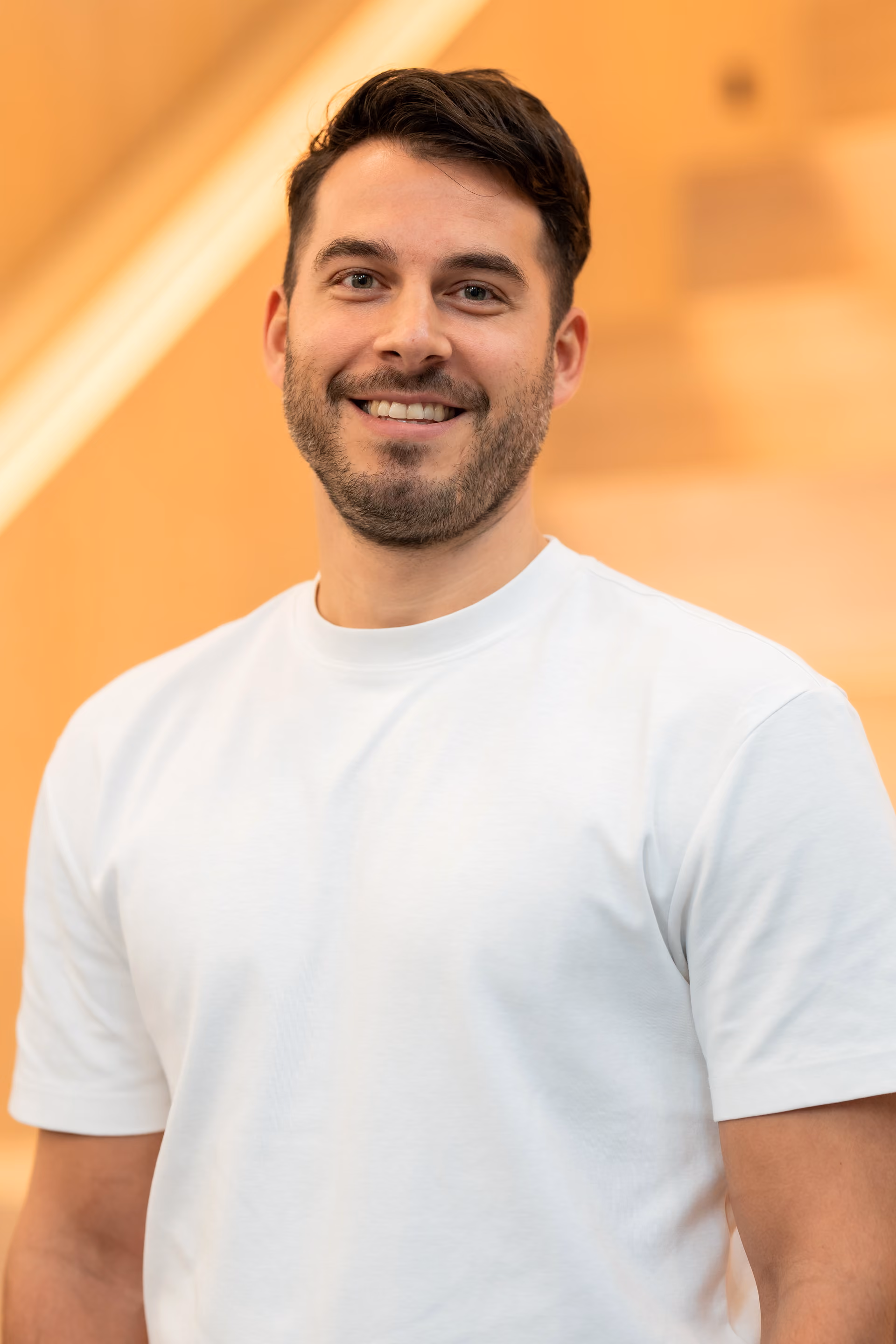 Lächelnder Mann mit dunklem Haar und Bart in einem weissen T-Shirt vor unscharfem beige Hintergrund.