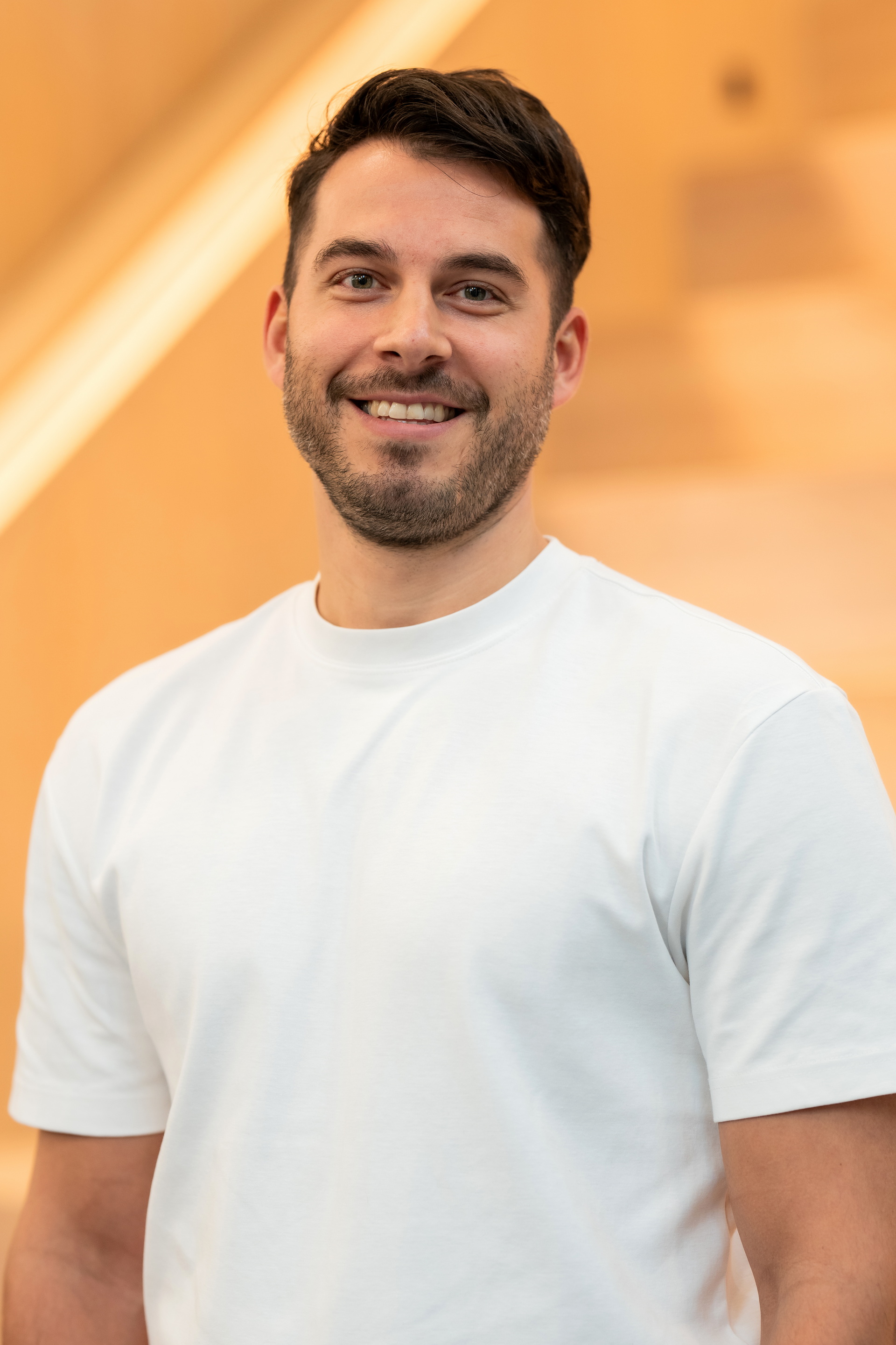 Lächelnder Mann mit dunklem Haar und Bart in einem weissen T-Shirt vor unscharfem beige Hintergrund.