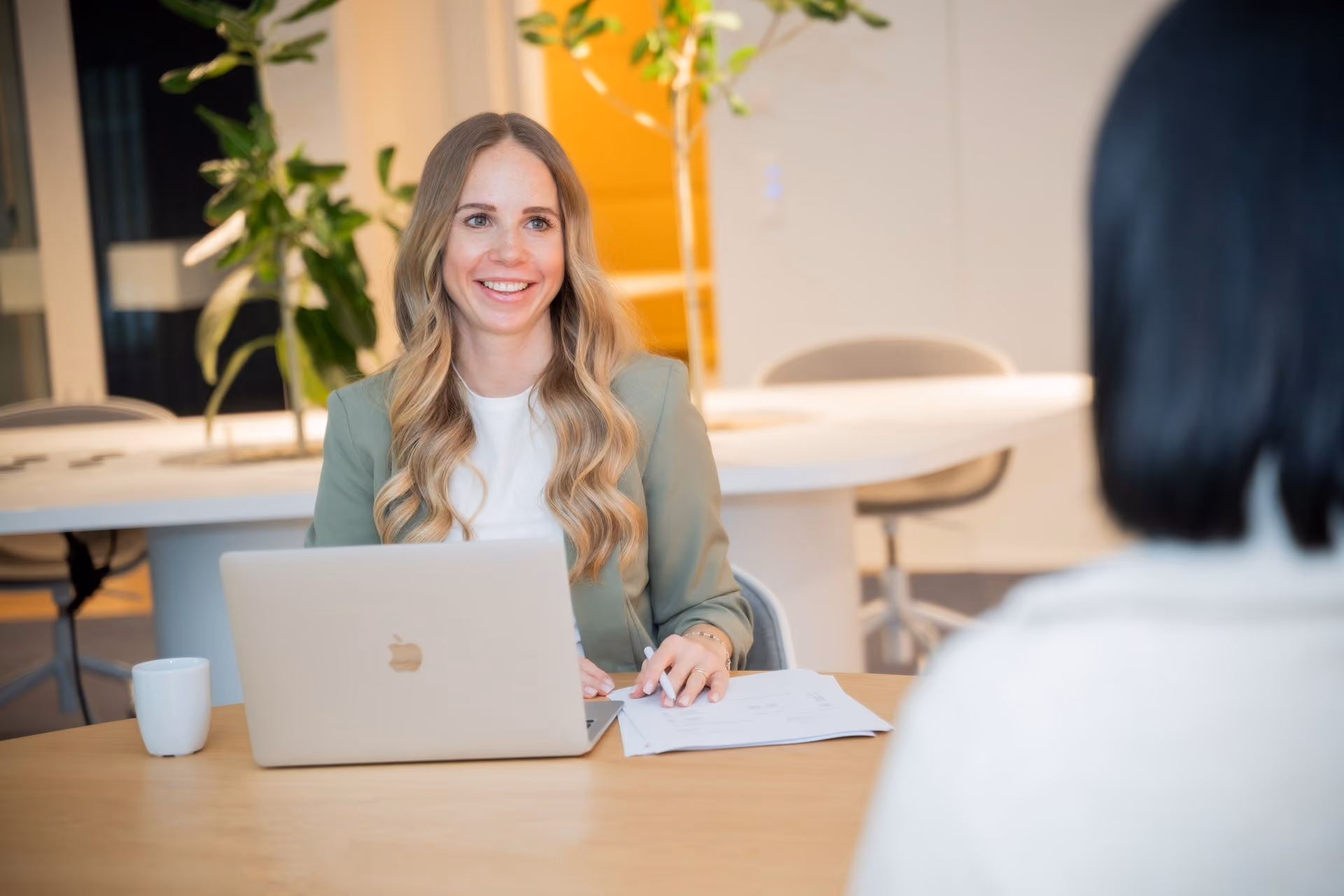 Lächelnde Frau mit langen blonden Haaren sitzt an einem Tisch mit Laptop und Unterlagen in einem modernen Büro - ein Symbol für Unternehmensberatung.