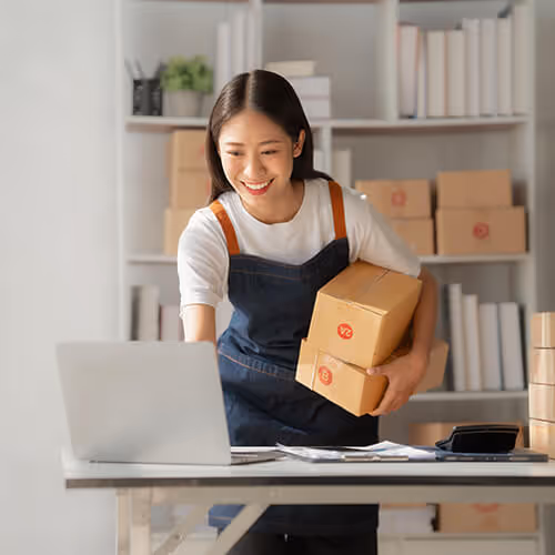 A woman holding packages whilst looking at a laptop