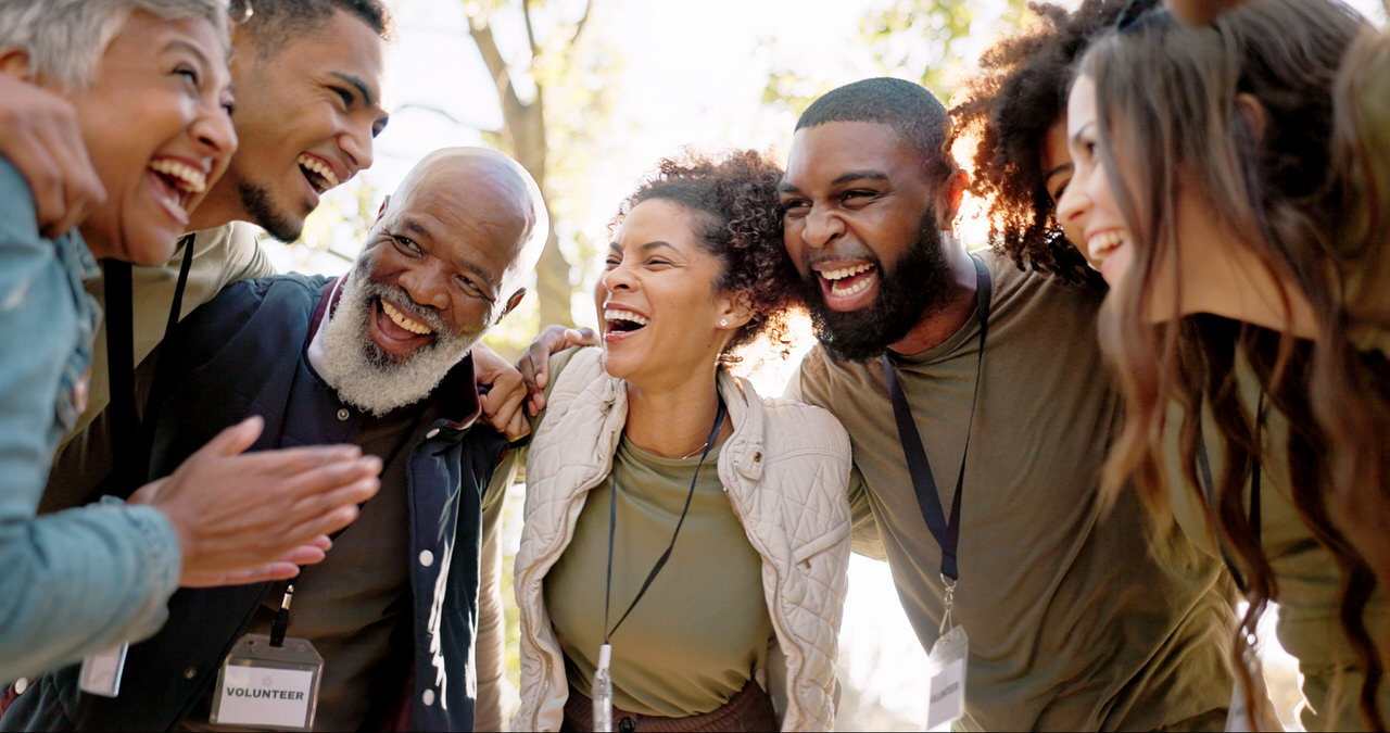 A group of cheerful volunteers standing close together outdoors, smiling and laughing with arms around each other, wearing volunteer badges.