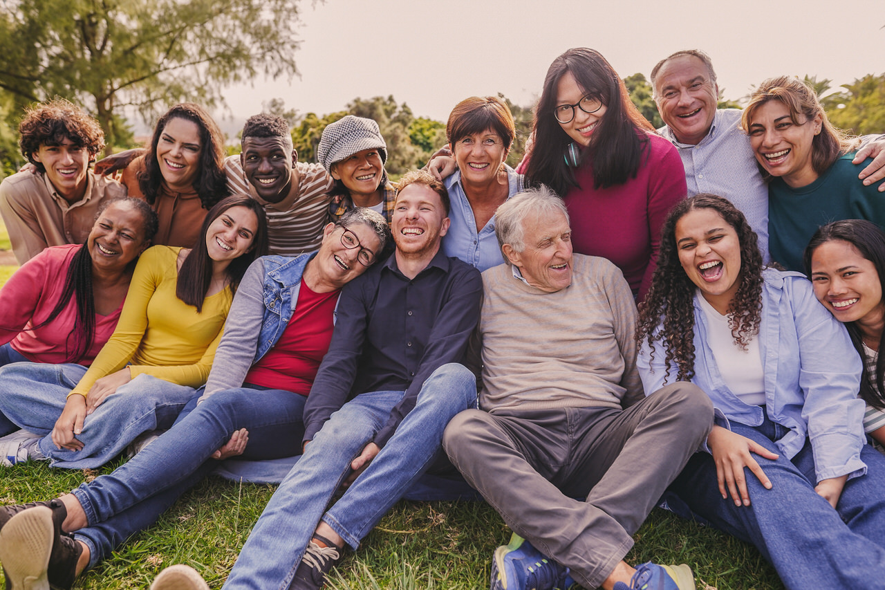 A diverse group of 15 people of various ages sitting closely together on grass outdoors, smiling and laughing in a joyful, friendly setting with trees in the background.
