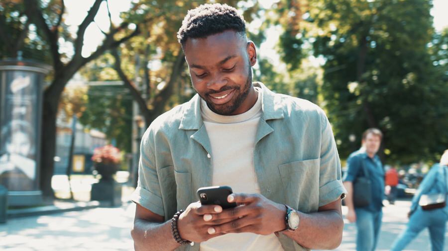 Smiling man wearing a light green shirt and watch, looking down at his smartphone outdoors in a sunny park with trees and people in the background.