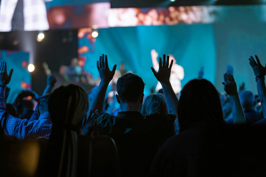 Audience raising hands in a dimly lit concert or event venue with blue lighting and a blurred stage in the background.