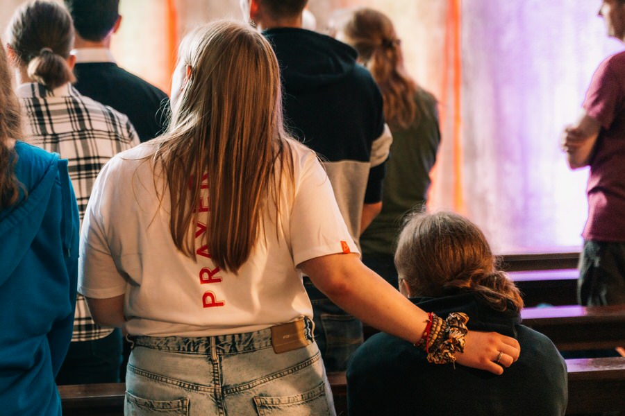People standing in a room with wooden benches, one person wearing a white shirt with 'PRAYER' in red on the back has their arm around another seated person.