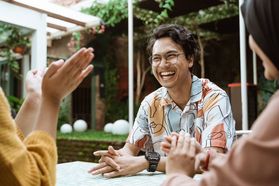 A young man with glasses and a colorful abstract patterned shirt laughing and sitting at a table outdoors, while two people across from him clap their hands.