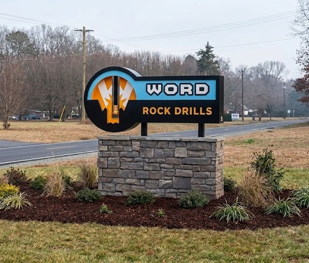 Stone base sign next to a road reading 'WORD ROCK DRILLS' with a drill bit logo.