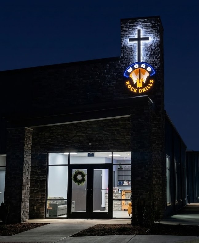 Building entrance at night with lit cross and Word Rock Drills sign on stone facade.