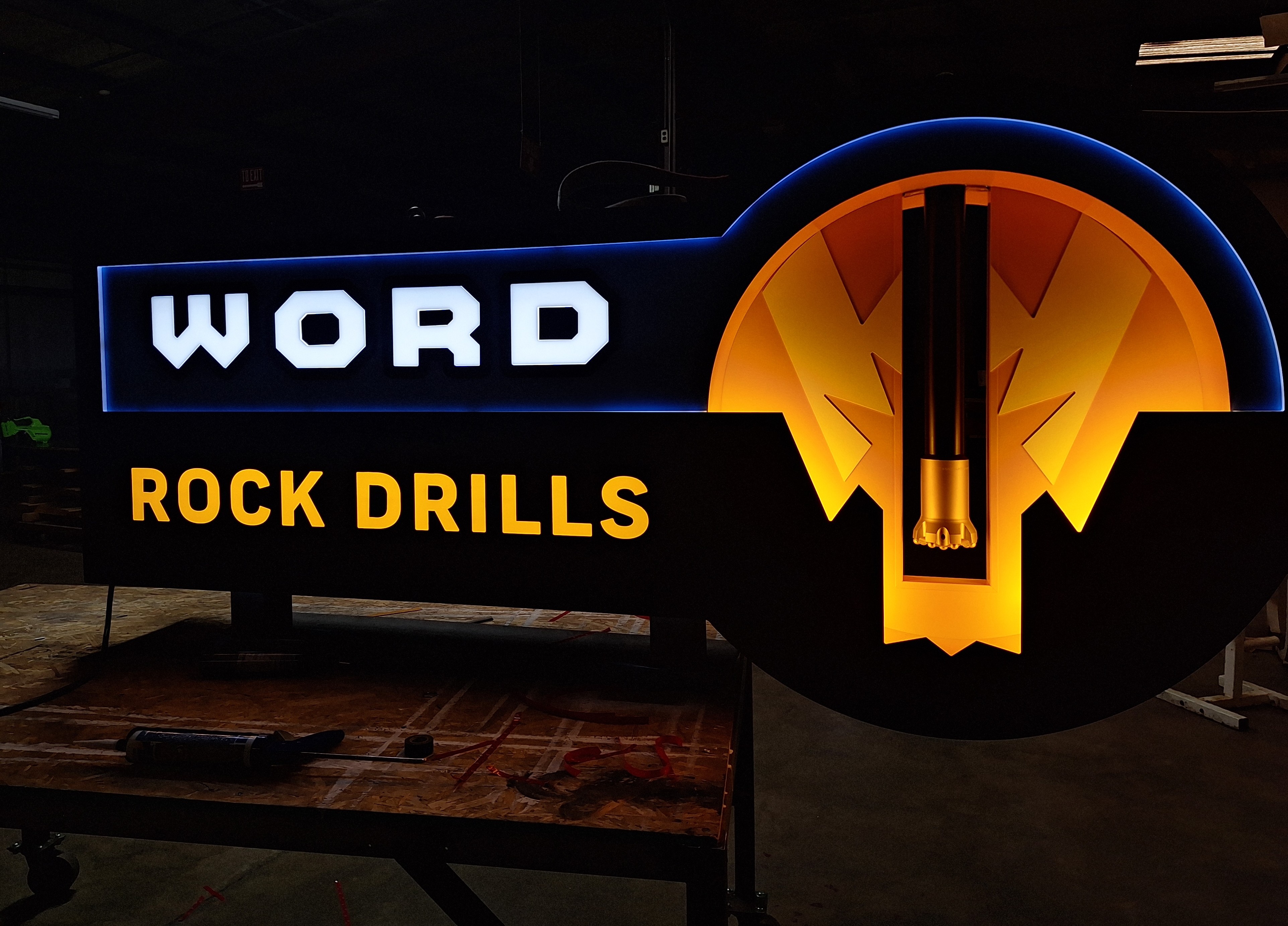 Illuminated sign displaying 'WORD ROCK DRILLS' with a rock drill bit graphic and glowing blue and yellow lights.