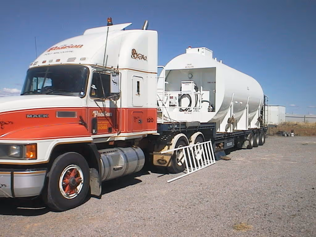 White and red Mack semi-truck with a large white cylindrical tank trailer parked on gravel under a clear blue sky.