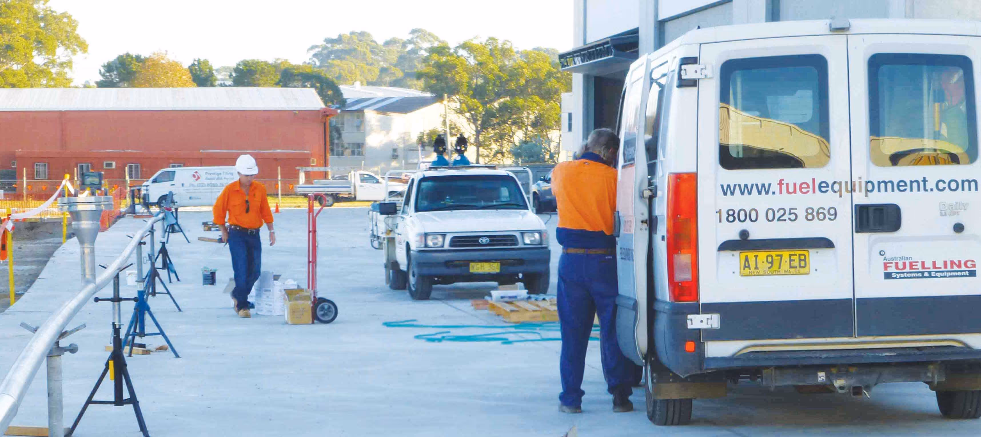 Two workers in orange shirts at a construction site with white utility vehicles and equipment.
