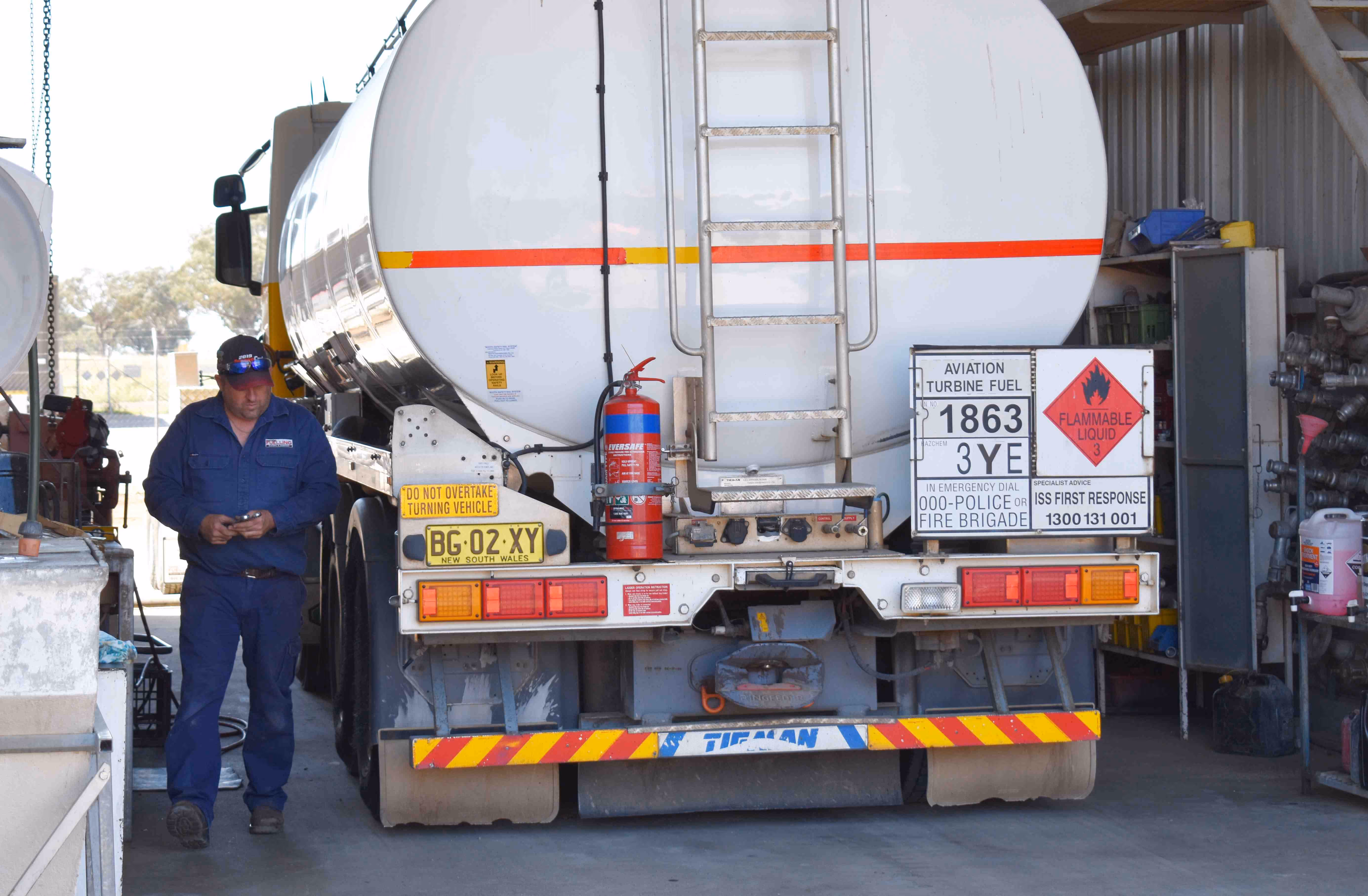 Rear view of a white aviation turbine fuel tanker truck with safety signs and a man in blue coveralls standing beside it.