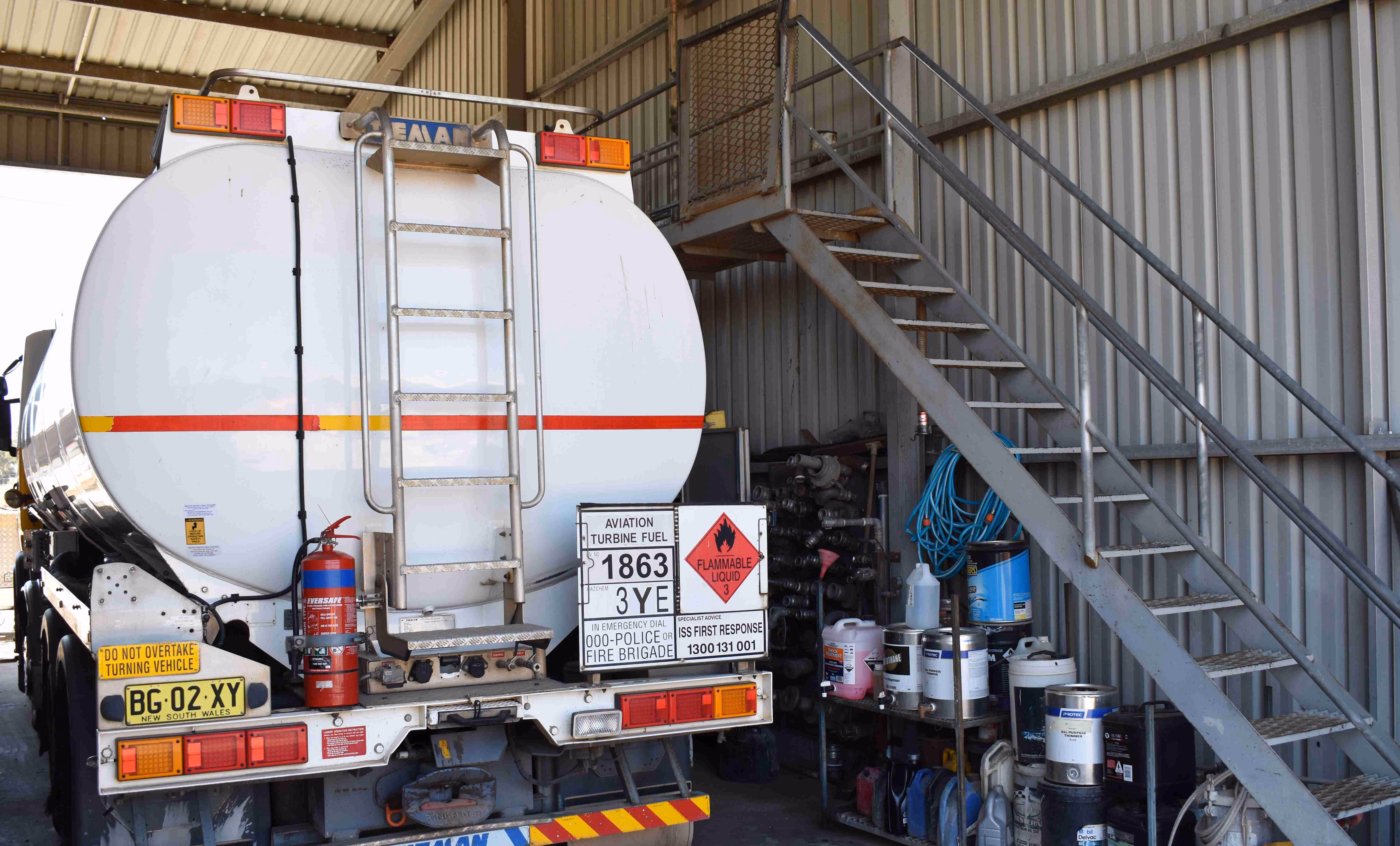 Rear view of a white aviation turbine fuel tanker truck with hazard signs, parked inside a metal shed next to a staircase and various fuel containers.