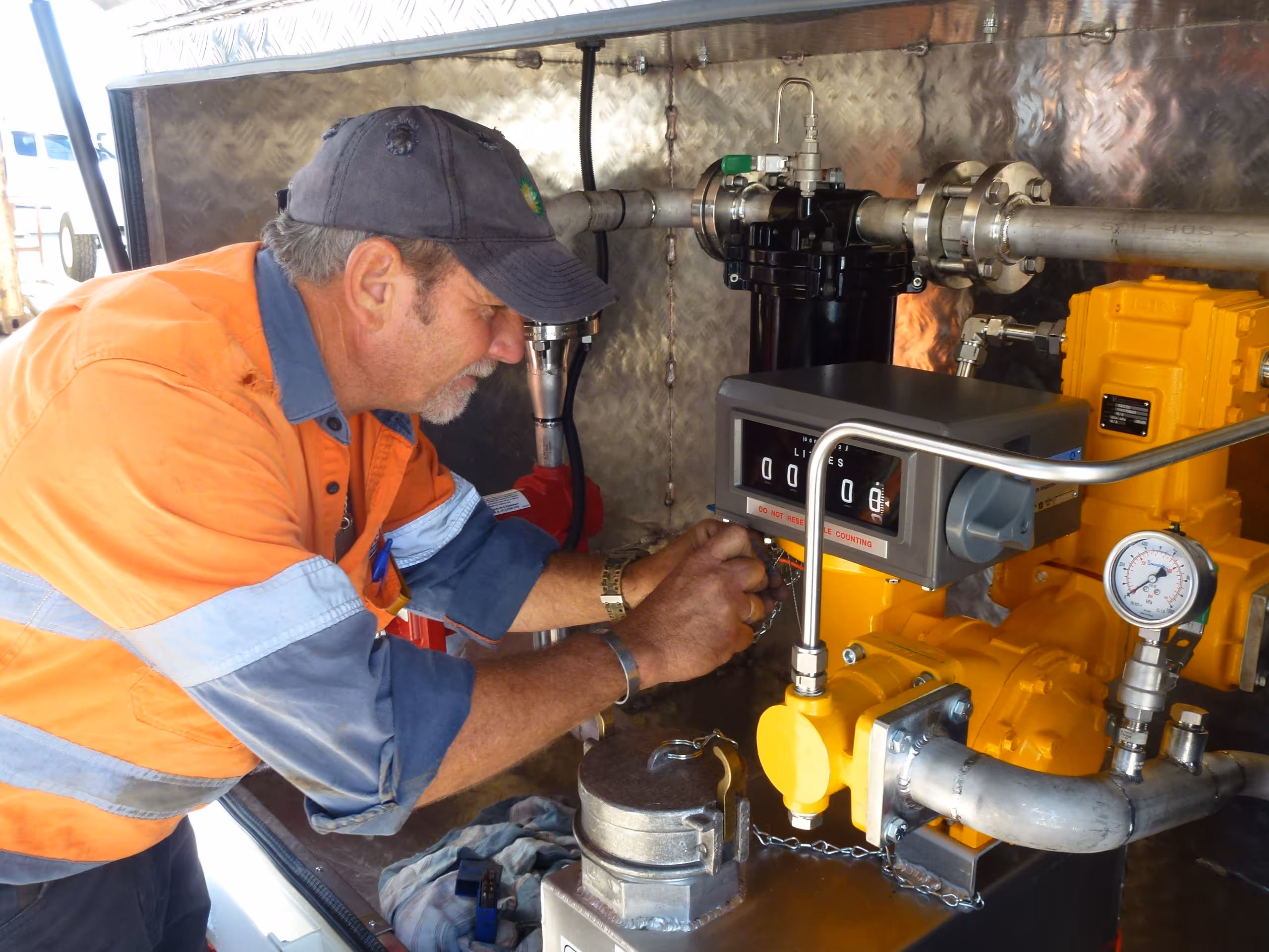 Technician in an orange safety shirt and cap working on a yellow industrial meter with pipes and a pressure gauge.
