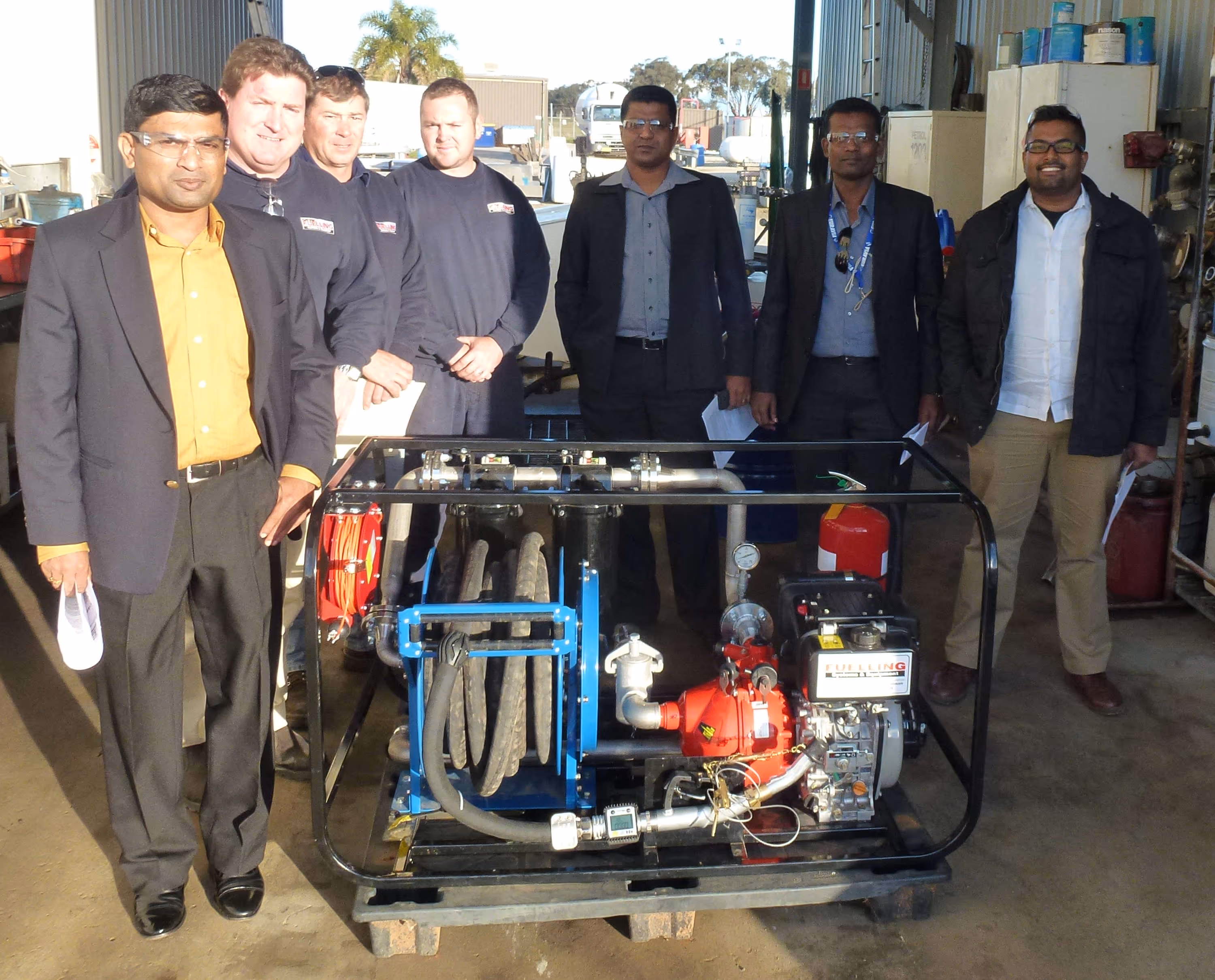 Seven men standing behind industrial equipment with hoses and pipes inside a workshop.