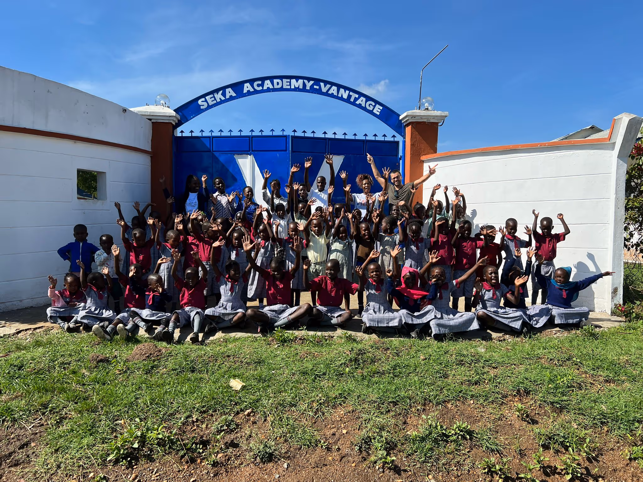 Pupils from Seka Academy Vantage, Kendu Bay, Lake Victoria, Kenya, waving as they sit outside the school gets with the school name above the gates behind them.