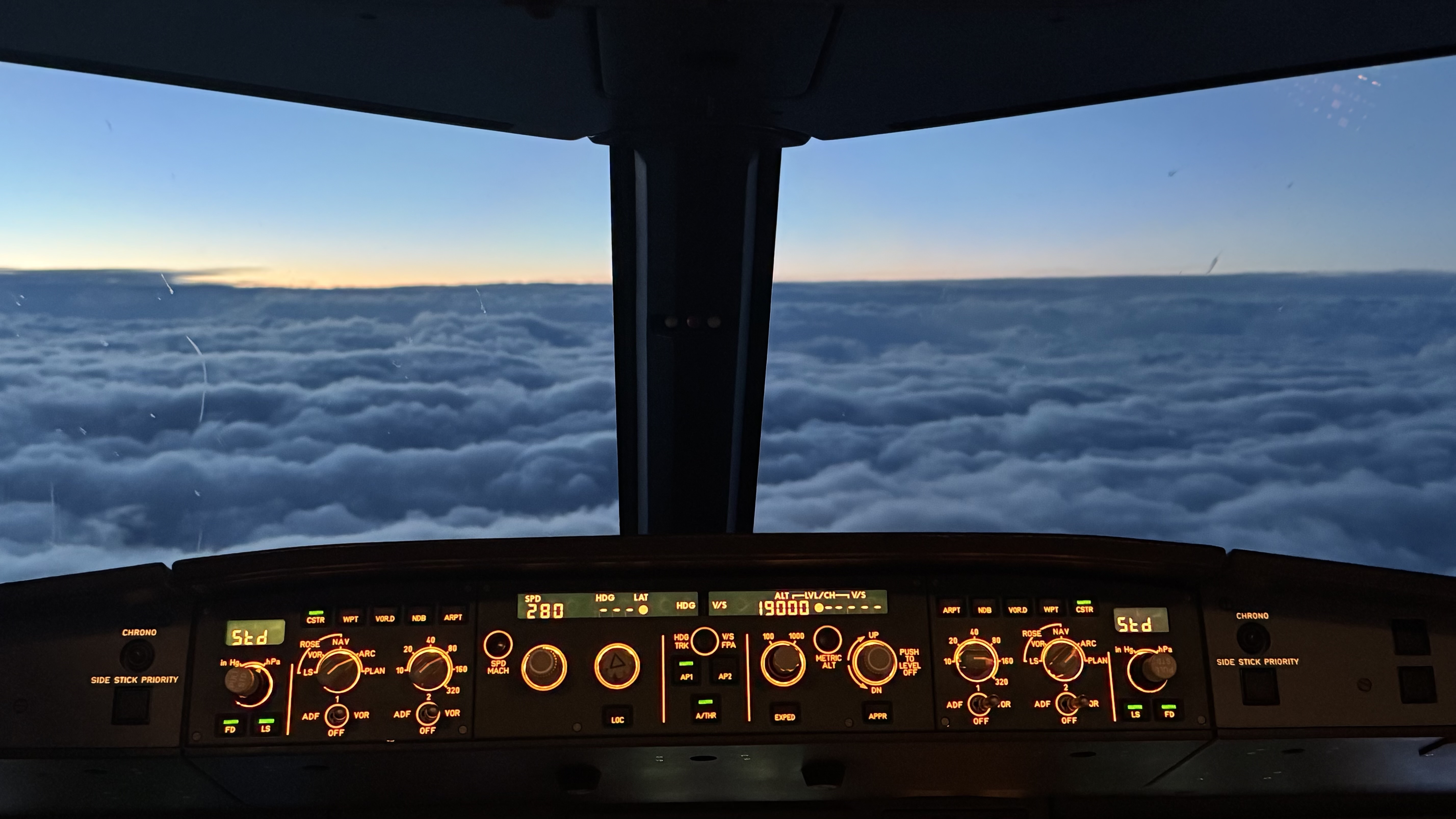 View from the cockpit with dials in the foreground. Clouds and a sunset are visible through the pilots windows.
