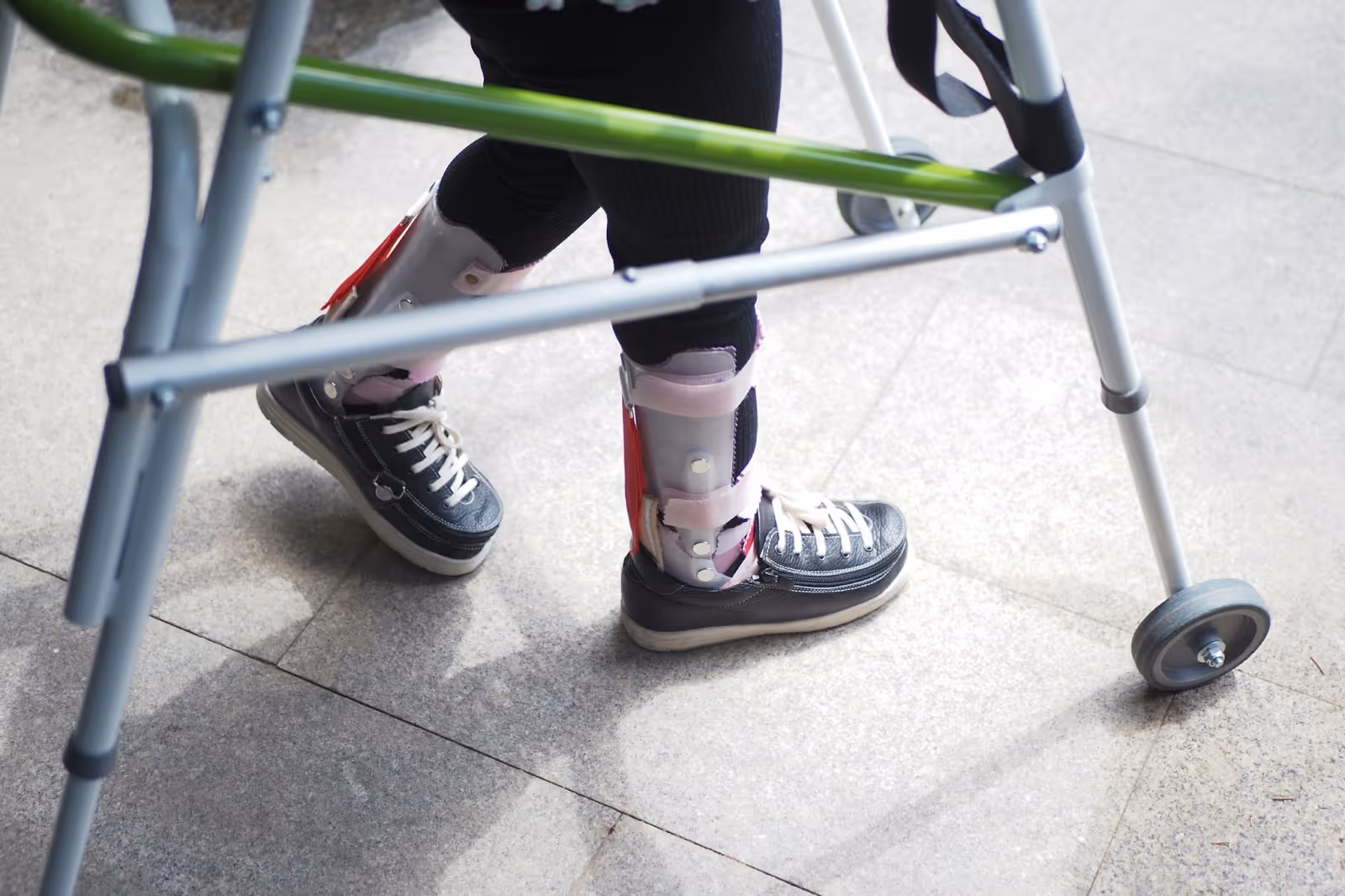 Person wearing leg braces and black sneakers using a walker on a tiled floor.