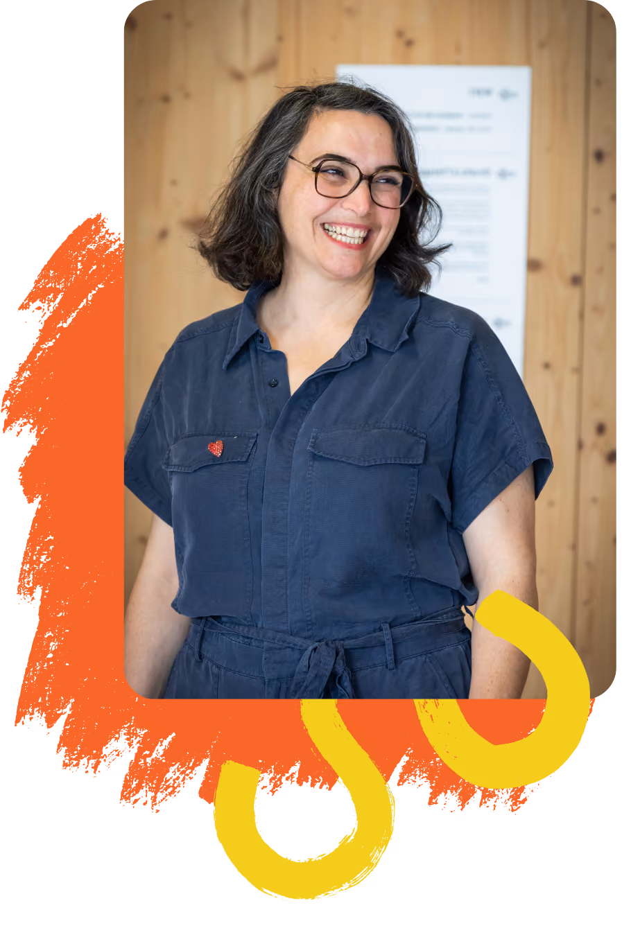 Louise Kolaï portant une combinaison bleue avec un petit badge en forme de cœur rouge sur la poitrine, cheveux mi-longs et lunettes, devant un mur en bois clair avec un panneau flou en arrière-plan.