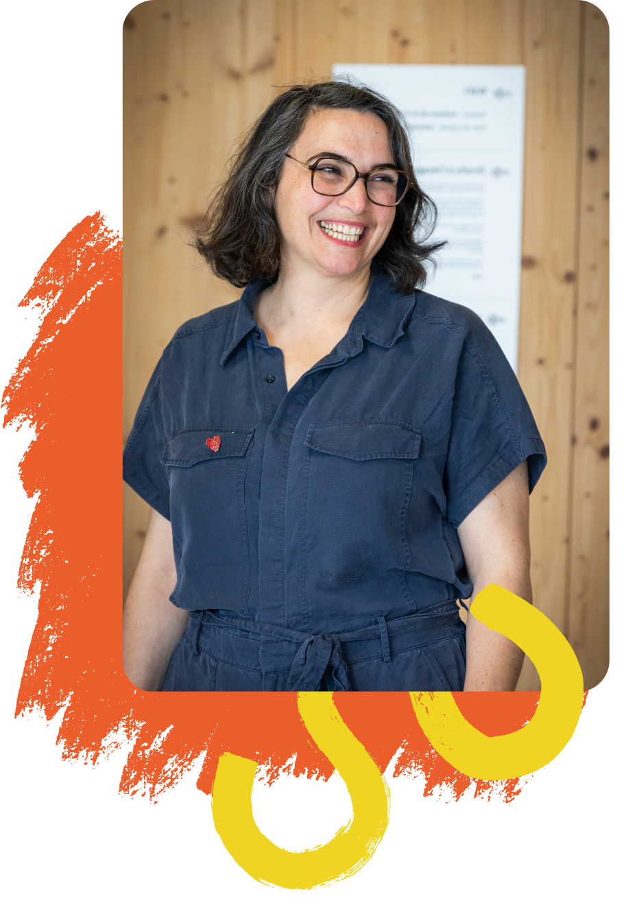 Louise Kolaï portant une combinaison bleue avec un petit badge en forme de cœur rouge sur la poitrine, cheveux mi-longs et lunettes, devant un mur en bois clair avec un panneau flou en arrière-plan.