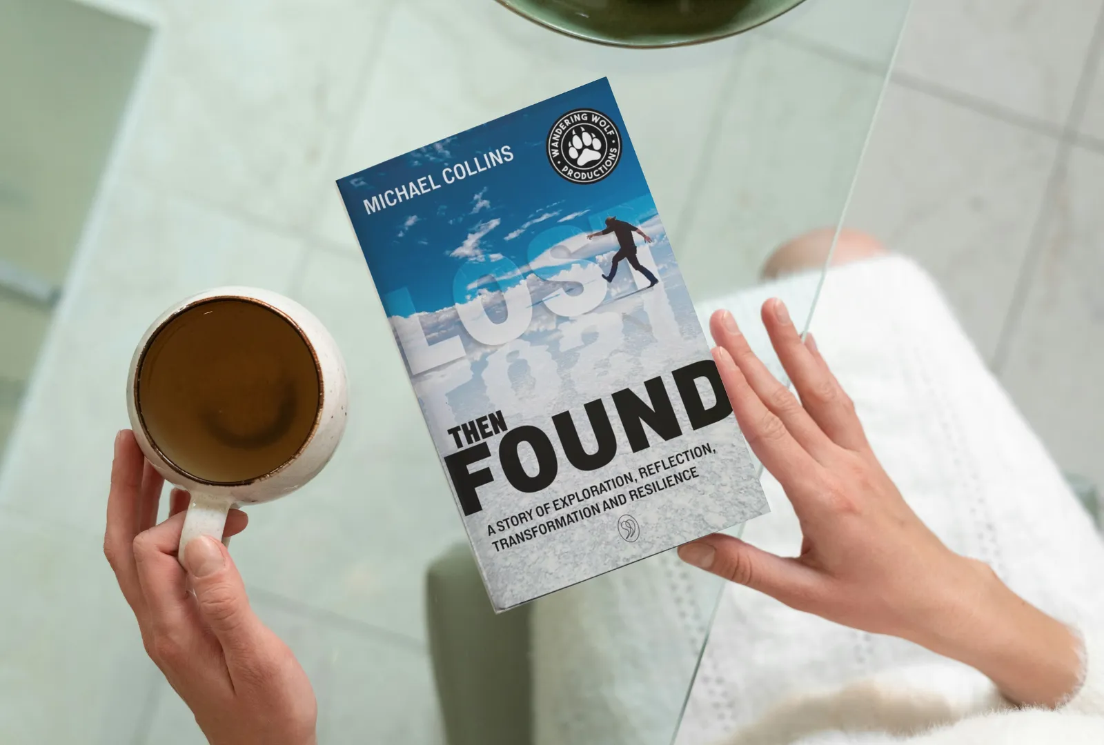 StoryTerrace - Books That Matter - Woman holding the book "Lost Then Found" by Michael Collins with a coffee at a glass table.