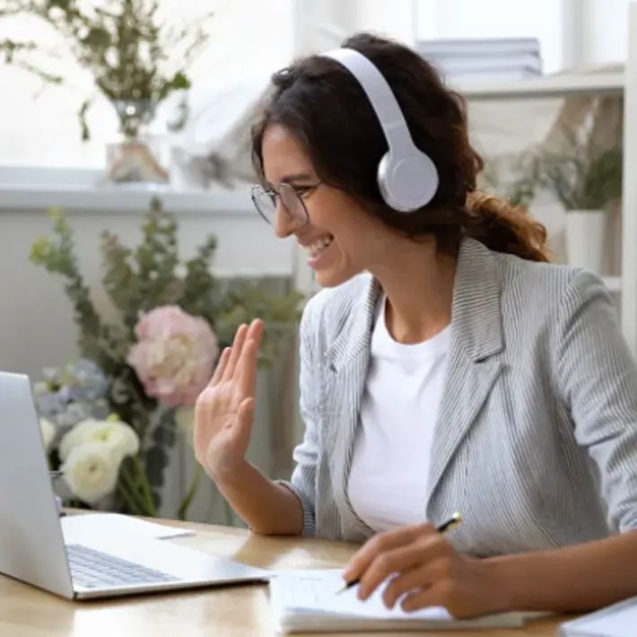 StoryTerrace - Books That Matter - Smiling woman wearing glasses and white headphones waving during a video call on laptop, taking notes.