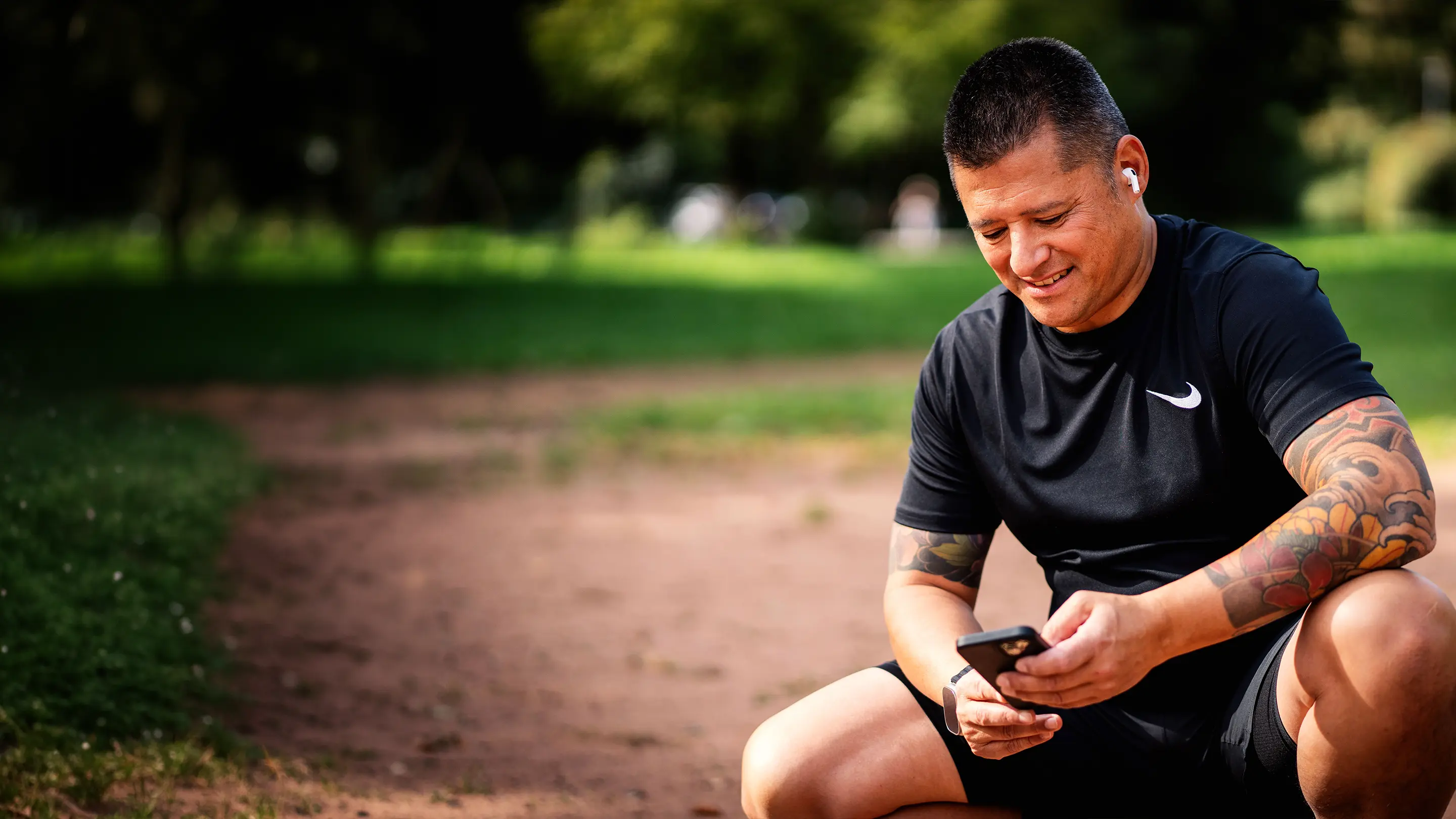 Sportlich gekleideter Mann mit Tätowierungen sitzt auf einem Parkweg, trägt kabellose Ohrhörer und schaut lächelnd auf sein Smartphone.