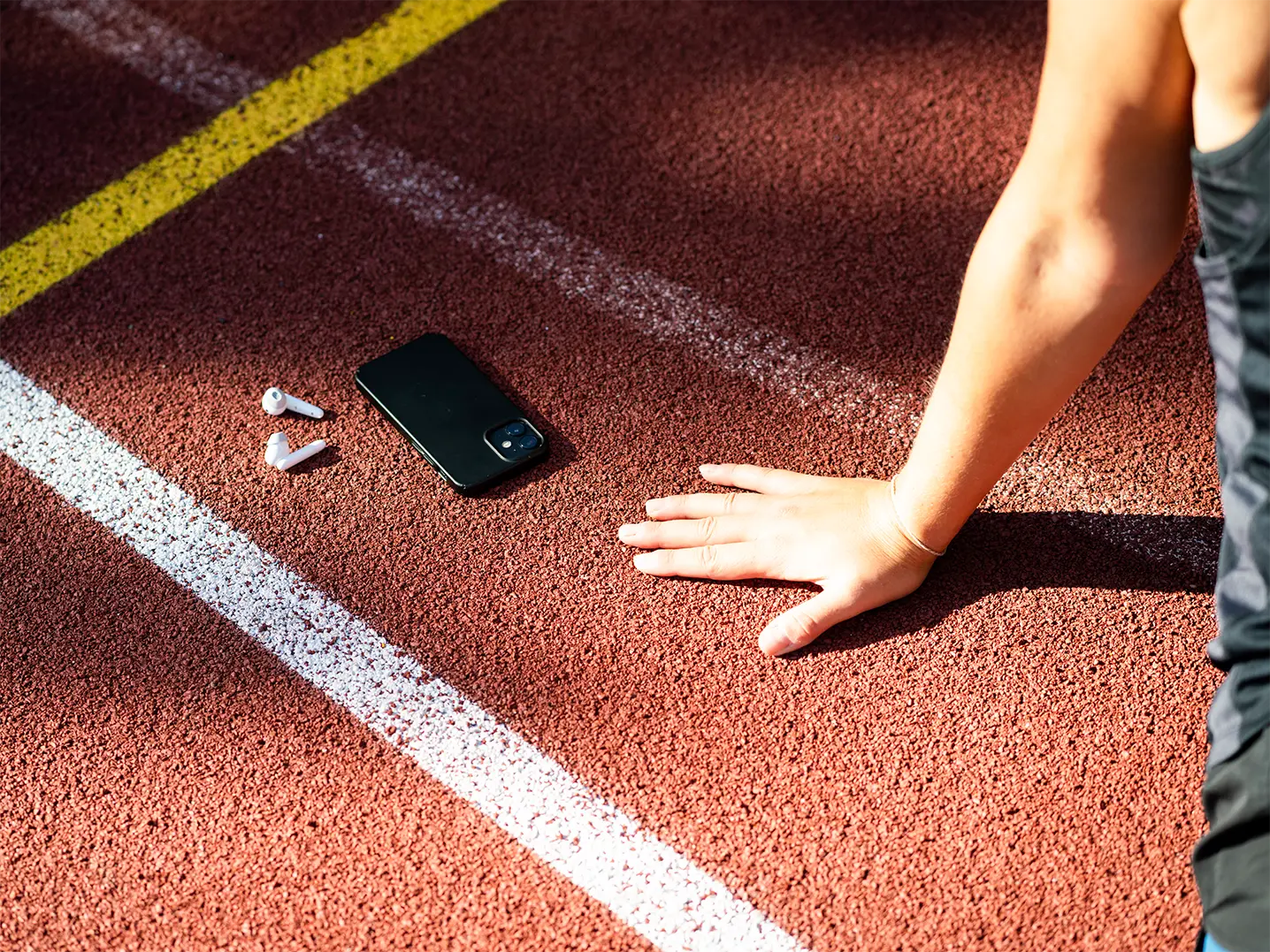 Hand einer sportlich gekleideten Person auf einer Laufbahn neben einem Smartphone und kabellosen Ohrhörern, mit sichtbaren weißen und gelben Markierungslinien.