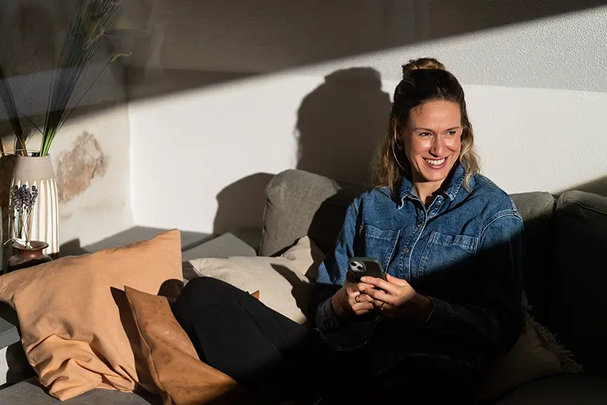 Lächelnde Frau sitzt auf einem Sofa im warmen Sonnenlicht und hält ein Smartphone in der Hand, neben ihr liegen mehrere Kissen.