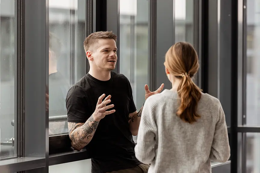 Zwei Personen stehen an großen Fenstern und unterhalten sich; ein Mann mit Tätowierungen gestikuliert beim Sprechen, während eine Frau mit Pferdeschanz ihm zugewandt zuhört.