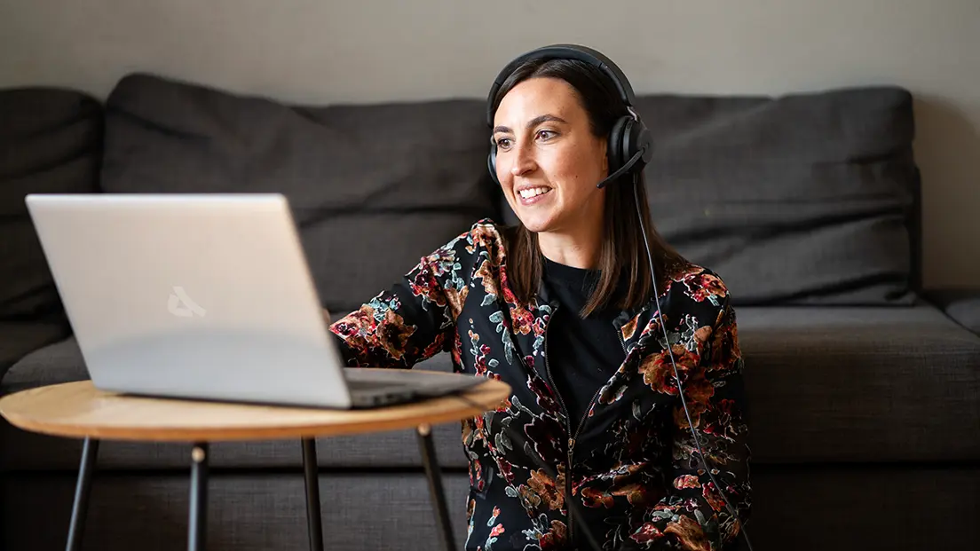 Frau mit Headset sitzt vor einem Laptop an einem kleinen Tisch und lächelt während eines Videoanrufs, im Hintergrund ein Sofa.